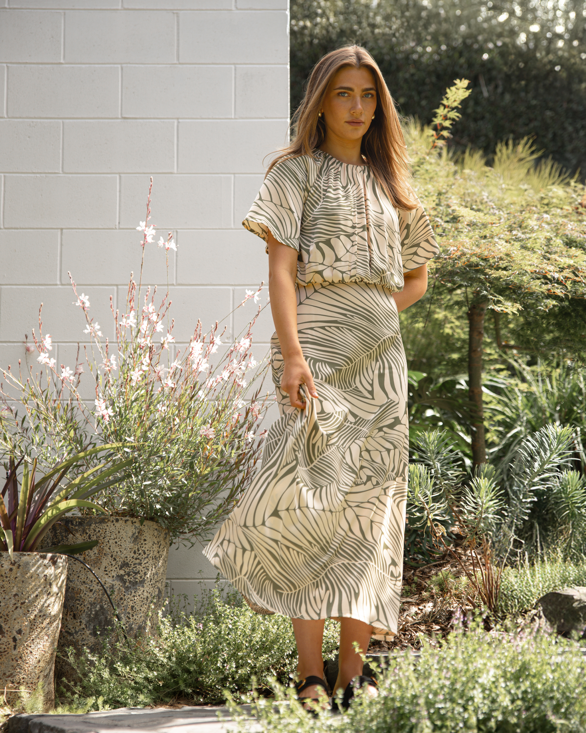 Woman in a patterned outfit standing in a garden with plants and a light-colored wall.