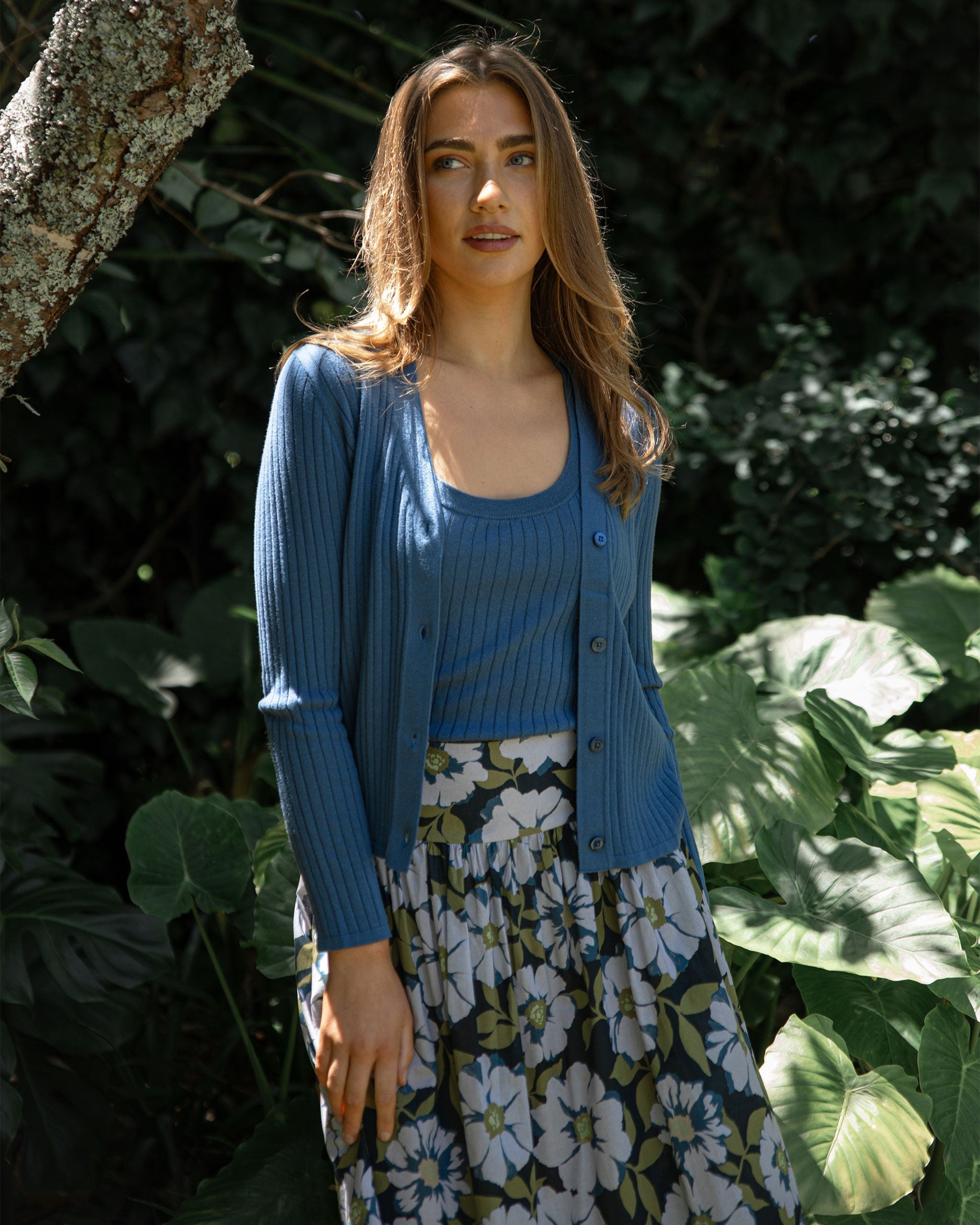 Woman wearing a blue cardigan and floral skirt standing among green foliage