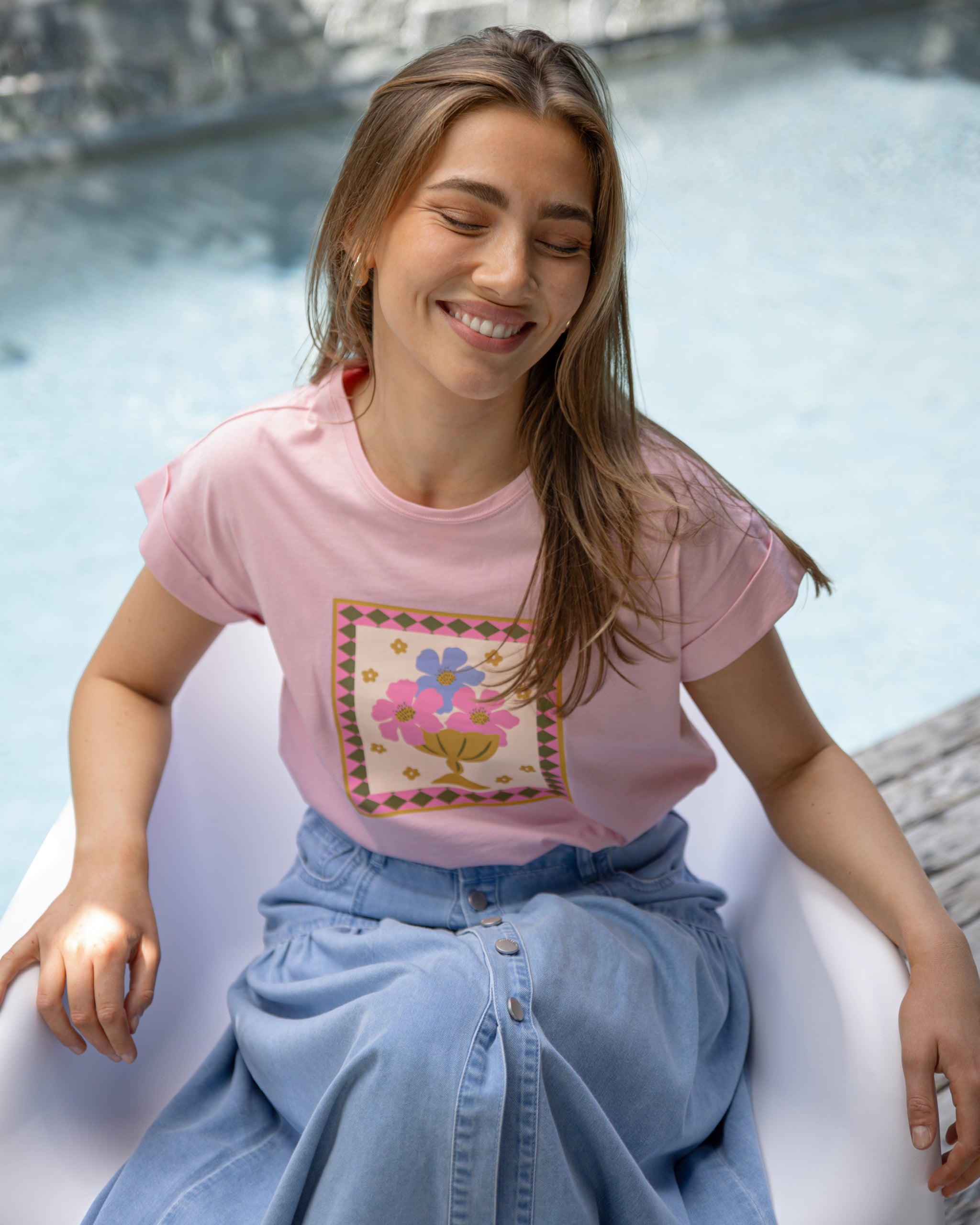 Woman wearing a pink t-shirt with a graphic design and blue jeans, sitting by a pool.
