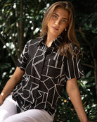 Woman wearing a black geometric patterned shirt sitting outdoors with greenery in the background
