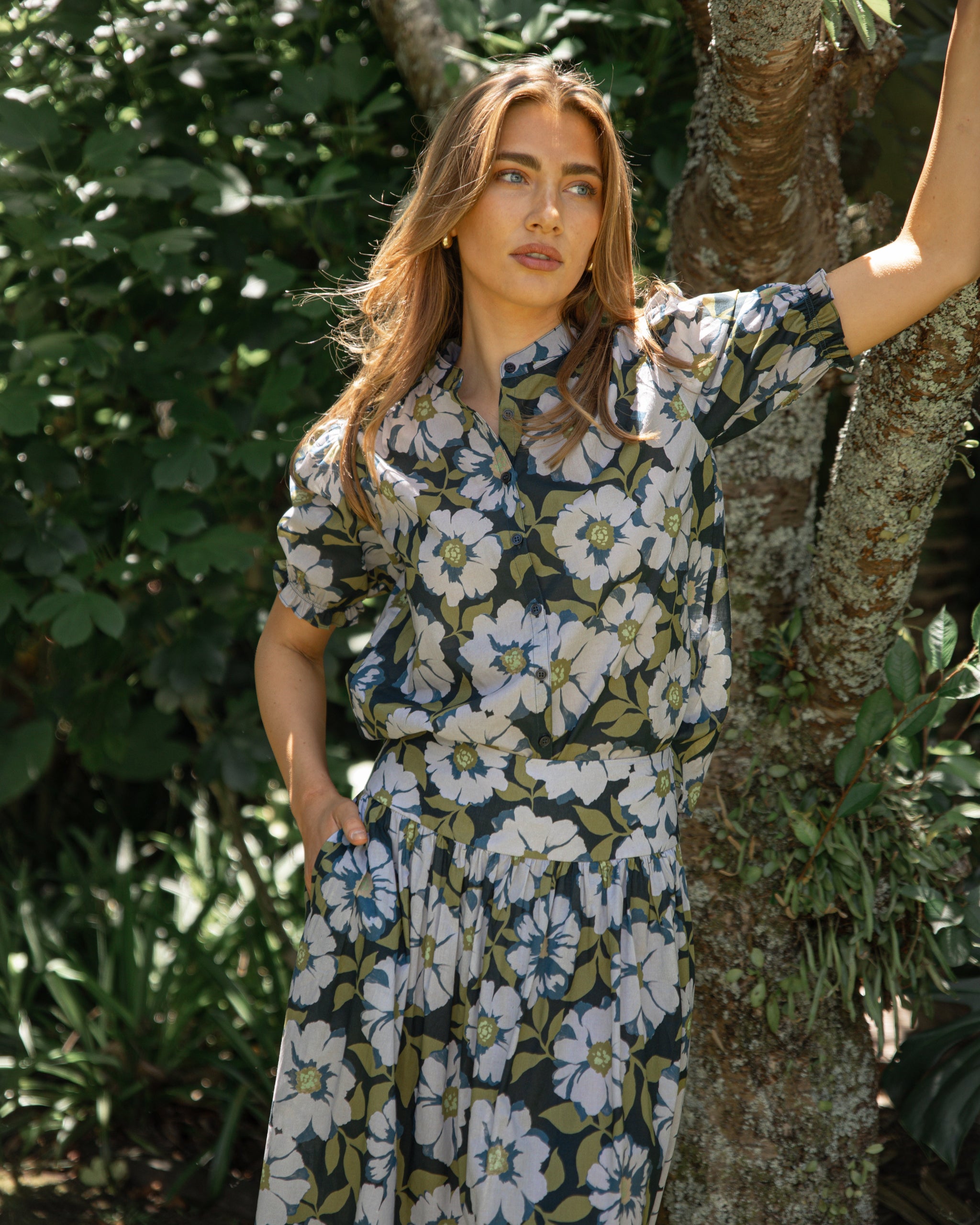 Woman in a floral dress standing next to a tree in a garden setting